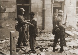 A disheveled man stands on a city street in front of a uniformed soldier surrounded by rubble. The soldier has his hand on the man’s shoulder and appears to be leading him. There are two other uniformed soldiers on the right side of the photograph. Both hold firearms.
