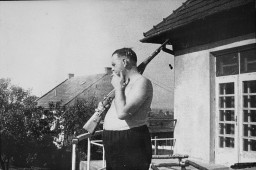 A man with short dark hair and light skin tone stands on a balcony while smoking and balancing a rifle on his shoulder. He appears to be wearing a white tank top and dark pants. Buildings can be seen in the background.