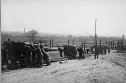 Standing with his arms behind his back, a kapo (right) oversees Jewish prisoners performing forced labor at the Płaszów camp. Kapos were concentration camp prisoners selected to oversee other prisoners on labor details. Płaszów, Poland, 1943–1944.