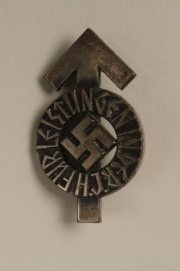 This Hitler Youth proficiency badge would have been awarded for the successful completion of a series of tests measuring physical and ideological proficiency. Success in these tests was rated according to criteria in the Hitler Youth identity document and performance book known as the Leistungsbuch. On this badge, the arrow shape (the tyr-rune) represents the warrior god Tyr. 
Beginning in 1933, the Hitler Youth and the League of German Girls had an important role to play in the new Nazi regime. Through these organizations, the Nazi regime planned to indoctrinate young people with Nazi ideology. This was part of the process of Nazifying German society. The aim of this process was to dismantle existing social structures and traditions. The Nazi youth groups were about imposing conformity. Youth throughout Germany wore the same uniforms, sang the same Nazi songs, and participated in similar activities. Badges such as this one emphasized the paramilitary nature of the Hitler Youth organization. It was designed to train boys as future fighters and soldiers for the Nazi cause. 