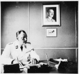 The commandant of Gross-Rosen, SS-Obersturmbannfuehrer Arthur Roedl, at his desk with a photograph of Adolf Hitler hanging on the wall. Gross-Rosen, Germany, 1941.