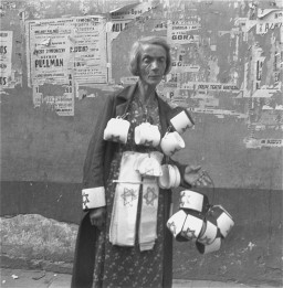 An emaciated woman with medium-length light hair and medium skin tone stands in front of a wall selling armbands with a six-pointed star. She is wearing a coat that has one of the bands around the end of the sleeve. She carries several that have already been sewn together in a loop in her left hand with more around her neck. She has several unsewn armbands hung from her waistband.