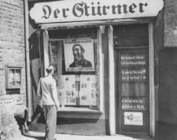 A young man looks at the antisemitic caricature in the display window of the Danzig office of "Der Stürmer." The poster reads: "The Jews are our misfortune." Danzig, 1939.
