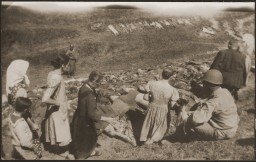 A line of people look towards a ditch filled with corpses. Uniformed soldiers observe the civilians.