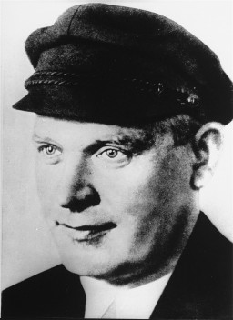 A black and white portrait of an adult man with light skin tone and short dark hair. He looks towards the left while wearing a dark cap and jacket.
