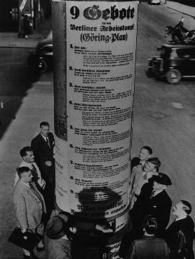 German pedestrians read Hermann Goering's "Nine Commandments for the Workers' Struggle,"  which included such exhortations as this one to German women: "take hold of the frying pan, dust pan and broom and marry a man."