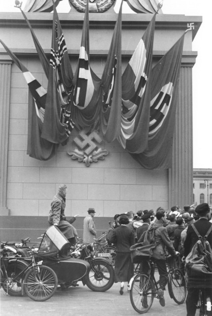 German spectators at a Nazi rally stand alongside a monument decorated with Nazi flags and a swastika emblem in Berlin. [LCID: 64450]