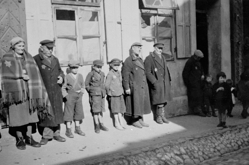 German Jewish adults and children wearing compulsory Jewish badges are lined up against a building.