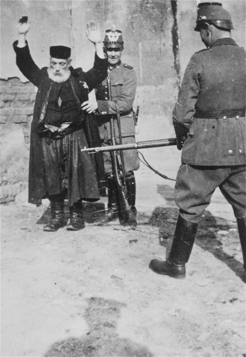 German policeman search an elderly, religious Jew at gunpoint in German-occupied Poland, circa 1941.
