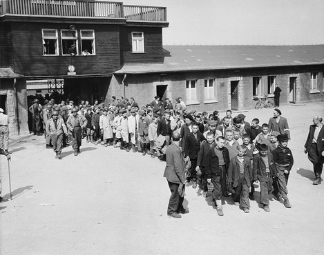 Escorted by American soldiers, child survivors of Buchenwald file out of the main gate of the camp.