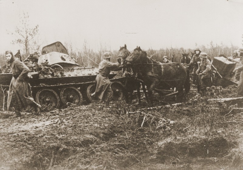 A German army column struggles through the mud, past a destroyed Soviet tank. [LCID: 09577]