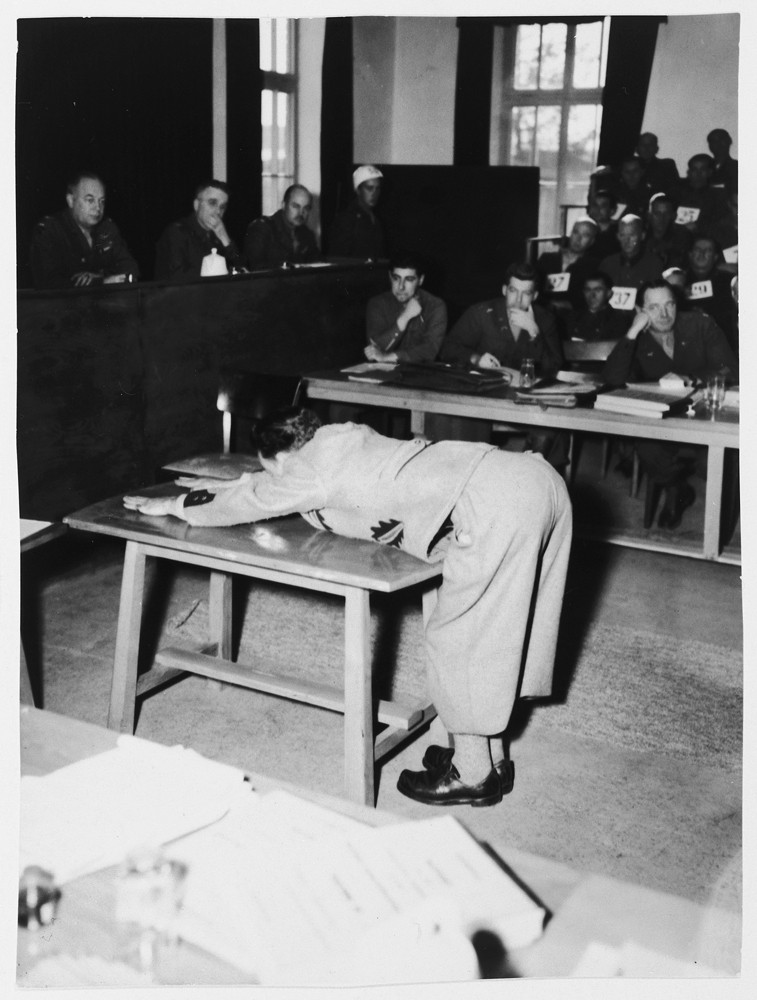 A prosecution witness demonstrates the position prisoners were forced to assume for punishment on the whipping block in the Dachau ... [LCID: 61085]