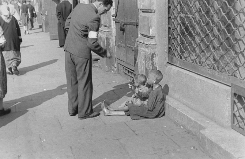 A Warsaw ghetto resident gives money to two children on a Warsaw ghetto street. [LCID: 89467]