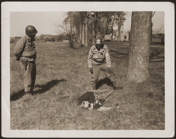An American medic and soldier stand by the corpse of a prisoner shot on the road near Gardelegen. [LCID: 28196]