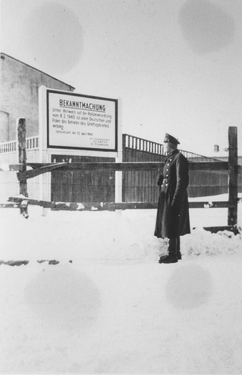 A member of the German Order Police Battalion 101 stands next to a sign marking the entrance to the Lodz ghetto in German-occupied Poland, 1940–1941. The German text of the sign, reads: "Announcement: In accordance with a police order of February 8, 1940, all Germans and Poles are forbidden entry into the ghetto area."