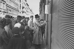 Onlookers watch as a Jewish man is forced to paint anti-Jewish graffiti on a shuttered storefront. Vienna, Austria, March 1938.