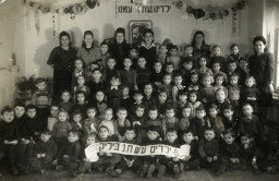 Bialik kindergarten students pose together in the Mariendorf displaced persons (DP) camp, circa 1946–1948. The children hold a banner that reads, "Ch, N. Bialik Kindergarten." A portrait of Theodor Herzl hangs on the back wall. Above the portrait is is a Hebrew banner that reads "Our children, the future of our nation."
Benjamin Markowicz is in the fourth row, second from the left.