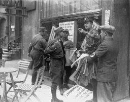在德国的某商业街上，一名纳粹冲锋队队员在指示其他人张贴反犹抵制标语的位置。一位戴着臂章的德国人手持一摞反犹标语，而冲锋队队员将它们贴到犹太人的商业街上。大多数标语都写道：“德国人，把你们自己从犹太人恶劣的宣传中解放出来，只从德国人自己的商店里购买物品”。拍摄地点：德国，拍摄时间：1933 年 4 月 1 日左右。