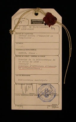 Evidence tag from the trial of Klaus Barbie in Lyon, France. This standard police form lists Barbie's infractions as crimes against humanity and complicity, concepts defined at the International Military Tribunal at Nuremberg decades earlier. The line in which the victims' names would be recorded is left blank. February 25, 1983.