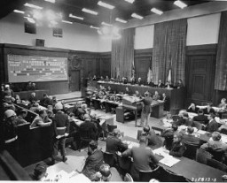 Le major américain Frank B. Wallis (debout, au centre), membre du personnel juridique du procès, présente l'accusation au Tribunal militaire international de Nuremberg. Un graphique (en haut à gauche) indique la position des accusés (en bas à gauche) dans l'organigramme du Parti nazi. À droite se trouvent les avocats des quatre pays ayant engagé des poursuites. 22 novembre 1945.