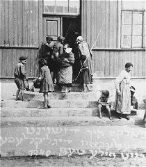 Mujeres y niños en la puerta de un comedor de beneficencia mantenido por el Comité Judío Estadounidense para la Distribución Conjunta. El texto en yidish dice: "Los afortunados con tazones llenos". Zelechow, Polonia, 1940.