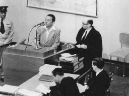 Henryk Ross testifies during Adolf Eichmann's trial. In addition to official duties as a photographer in the Department of Statistics in the Lodz ghetto, Ross secretly photographed scenes in the ghetto. To Ross' right is chief prosecutor Gideon Hausner, who holds some of Ross' photographs submitted as evidence. Jerusalem, Israel, May 2, 1961.