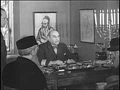 ドイツ軍は1939年9月にワルシャワに侵入し、その翌月、この町にユダヤ人評議会の設立を命じました。 ドイツ軍は、ワルシャワの旧ユダヤ人コミュニティ評議会のメンバーであったアダム・チェルニャクフを議長に選びました。 ここではドイツのニュース映画制作のために、ドイツの宣伝会社がチェルニャクフとゲットーからの請願者たちとの会合を仕立てています。 ドイツ側は、強制労働の要求とユダヤ人所有物の没収を含むドイツの命令をチェルニャクフが遂行することを予期していました。 チェルニクフ自身は、ドイツ側の措置の残忍性を緩和し、食料配給所、ワークショップ、および職業学校の設立を求めました。 彼は根強く条件の改善を懇願しました。 チェルニクフは、移送の対象となったユダヤ人の一斉検挙に協力するというドイツ側の要求に従う代わりに、1942年7月に自殺を選んだのです。