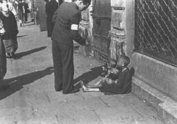 A Warsaw ghetto resident gives money to two children on a Warsaw ghetto street. Warsaw, Poland, between October 1940 and April 1943.