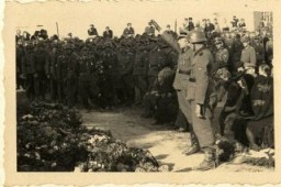SS officer Karl Höcker salutes in front of an array of wreaths during a military funeral near Auschwitz. The original caption for the photograph reads "Beisetzung von SS Kameraden nach einem Terrorangriff." (Burying our SS comrades from a terror attack.) Pictured in the background are Josef Kramer and Karl Moeckel.This image shows the aftermath of the September 13, 1944, bombing of IG Farben in which 15 SS men died in the SS residential blocks and 28 were seriously wounded. 
 
 