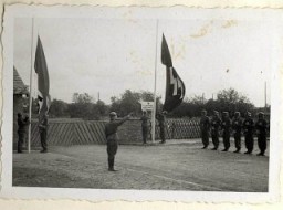The beginning of a ceremony to dedicate a new SS hospital in Auschwitz-Birkenau. A Nazi soldier salutes as the Nazi and SS flags are raised while a line of troops stand with rifles at attention during the dedication.
From Karl Höcker's photograph album, which includes both documentation of official visits and ceremonies at Auschwitz as well as more personal photographs depicting the many social activities that he and other members of the Auschwitz camp staff enjoyed. These rare images show Nazis singing, hunting, and even trimming a Christmas tree. They provide a chilling contrast to the photographs of thousands of Hungarian Jews deported to Auschwitz at the same time. 