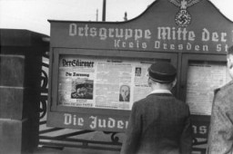 Remaja Jerman sedang membaca keluaran surat kabar Der Stuermer yang ditempel di papan pengumuman di pintu masuk kantor pusat partai Nazi di wilayah Dresden. Semboyan negara Jerman (kabur sebagian) di bagian bawah papan pengumuman berbunyi, "Yahudi adalah pembawa sial".