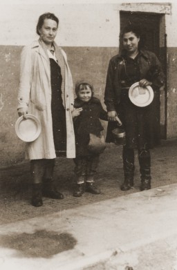 Two women and a child stand with metal bowls in front of a soup kitchen in the Cremona displaced persons (DP) camp in Italy, 1945. Pictured are Zelda Leikach and her daughter, Masha, with their friend Hinda.