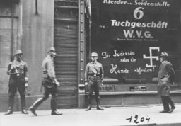 Peu après l’annexion de l’Autriche par les Allemands (l’Anschluss), des troupes d’assaut nazies montent la garde à l’extérieur d’un commerce détenu par un Juif. Un graffiti peint sur la fenêtre déclare : “Toi espèce de porc juif, que tes mains pourrissent!” Vienne, Autriche, mars 1938.