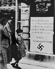 Viennese pedestrians view a large Nazi sign posted on a restaurant window informing the public that this business is run by an organization of the Nazi Party and that Jews are not welcome. Vienna, Austria, March-April 1938.