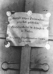 O cartaz diz:  "Empresa fechada pela Polícia devido a atividades especulativas.  O proprietário encontra-se sob custódia protegida em Dachau".  Assinado pelo Chefe de Polícia Heinrich Himmler. Munique, Alemanha, Abril ou Maio de 1933.