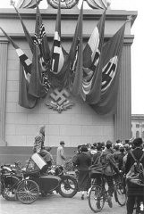Espectadores alemães de um comício nazista parados ao lado de um monumento decorado com bandeiras nazistas e o emblema da suástica. Berlim, Alemanha. 1937.