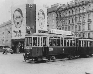 Un tramway décoré de croix gammées passe devant des panneaux d'affichage exhibant le visage d'Hitler.